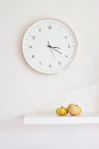 un grande orologio appeso ad parete bianca, sotto il quale su una mensola poggiano due piccole zucche gialle